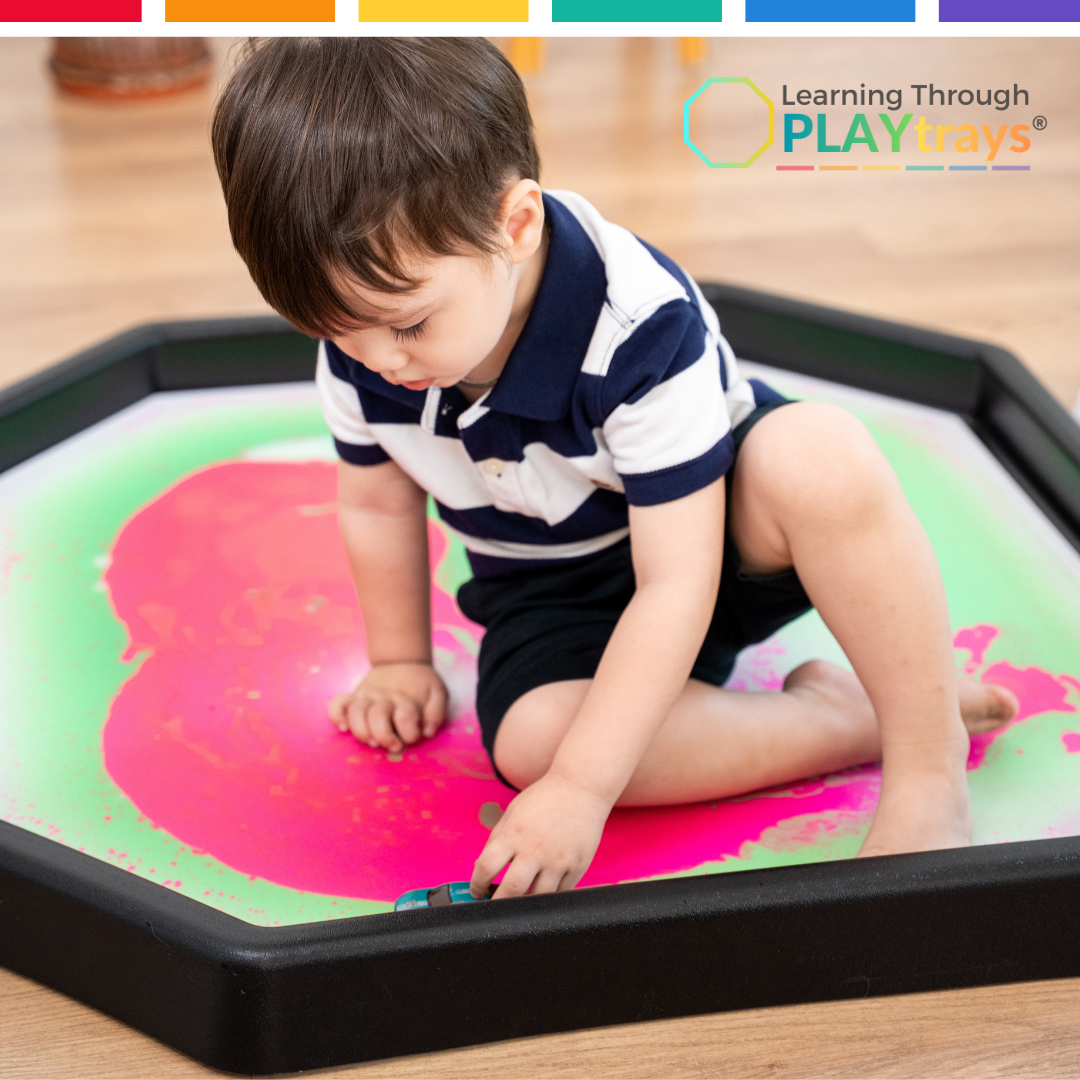 Child playing with a colorful tray on a wooden floor, featuring 'Learning Through PLAYtrays' logo.