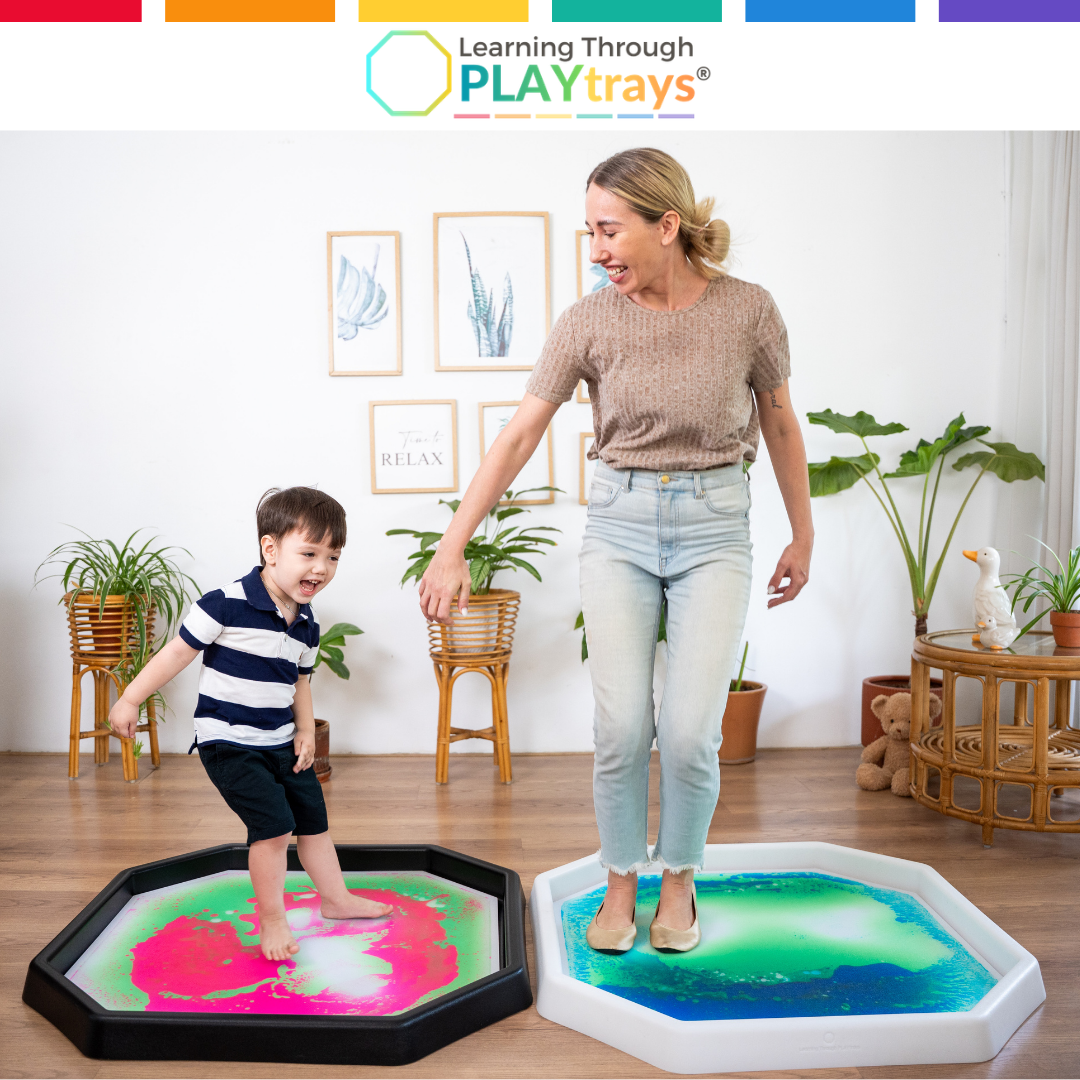 Woman and child playing on colorful play trays in a room with plants and furniture.