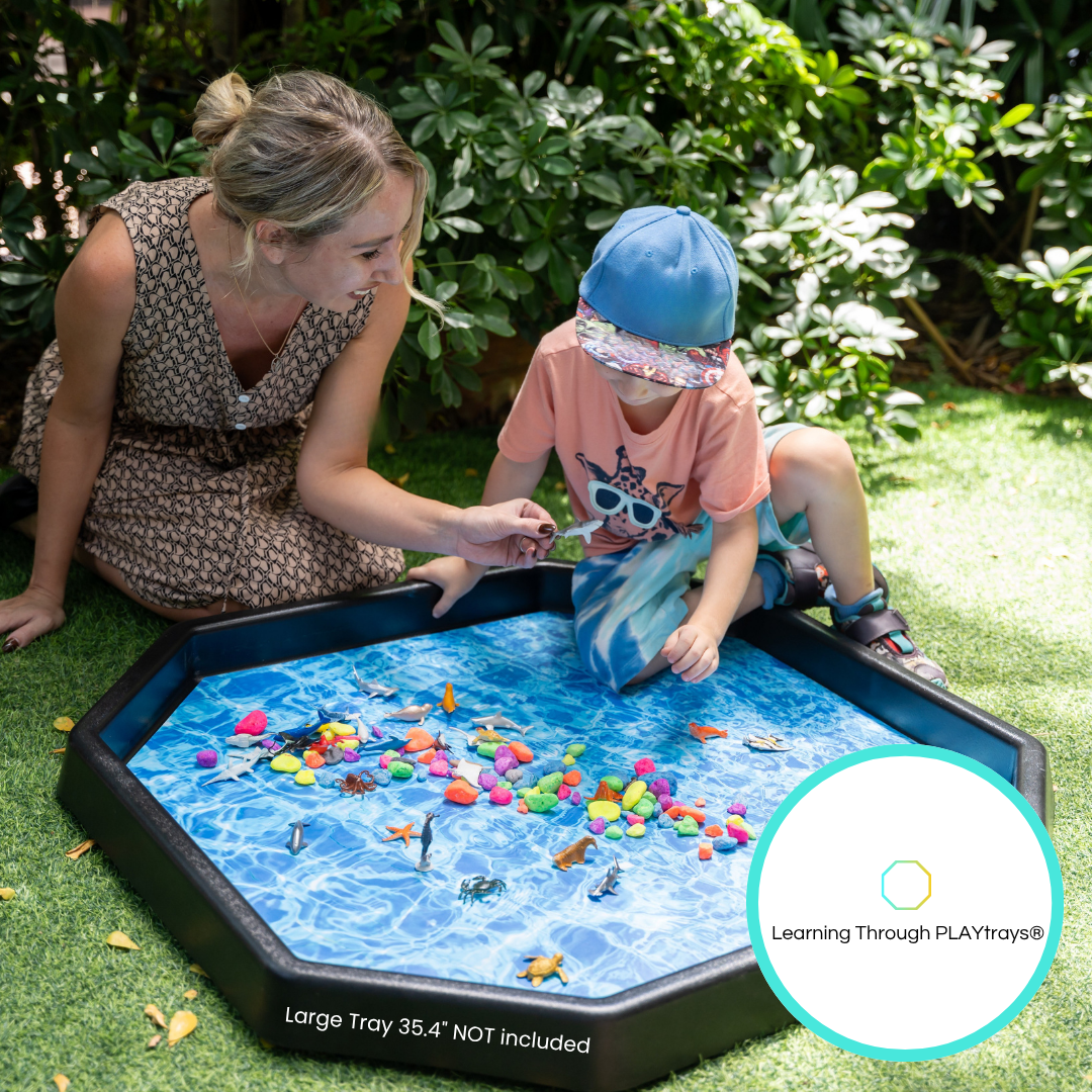 Child and woman playing with large blue water-themed Play Mat Insert inside a black octagonal tray outdoors on grass.