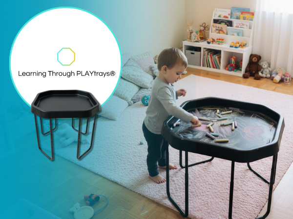 Child playing with a black tuff tray and chalk in a room with books and toys.